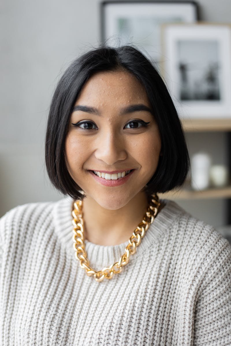 Portrait of a cheerful young woman wearing a gold necklace and sweater indoors with a blurred background.